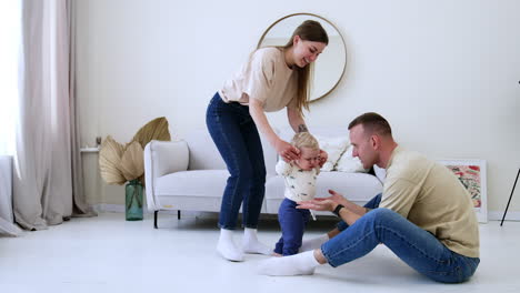 mother leads her lovely infant son by the room. father sits on the floor calling up his baby.