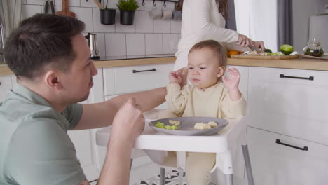 Father-Feeding-His-Happy-Baby-Girl-Sitting-In-Her-High-Chair-In-The-Kitchen-While-Mother-Cutting-Fruit-Behind-Them