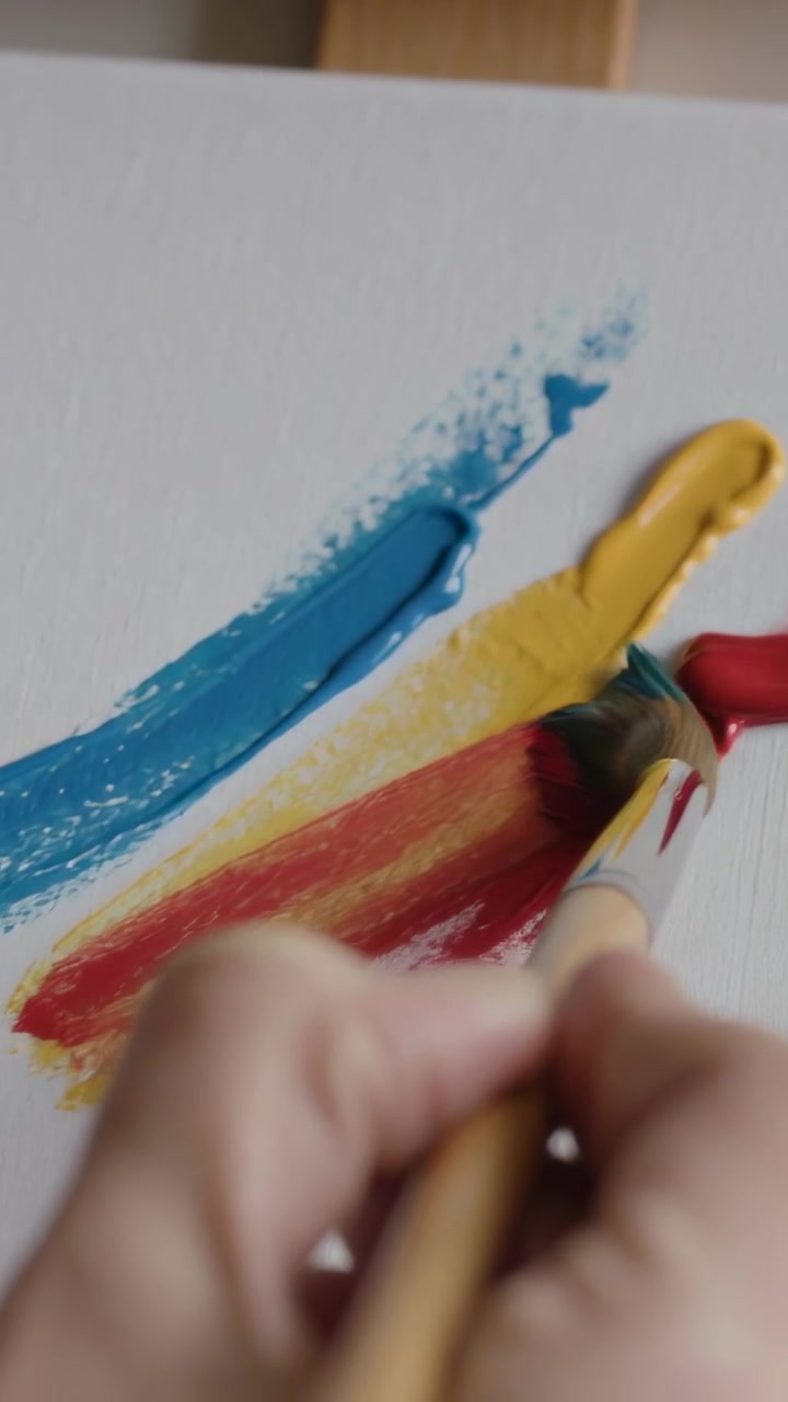 Video verticale: La mano di un pittore entra nell'inquadratura, sfumando rosso, giallo e blu con un pennello a ventaglio su una tela in studio.
