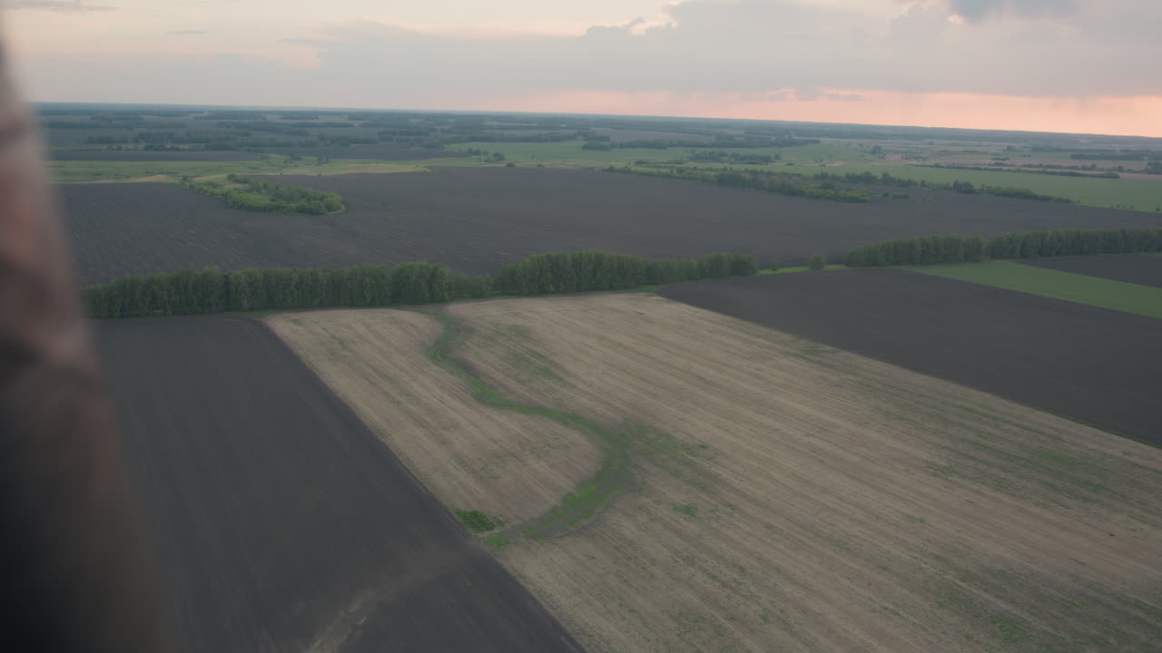 overhead aerial farmland patchwork of plowed and grassy fields under pastel sunset sky shot from hot air balloon basket revealing tranquil rural landscape textures and horizon glow