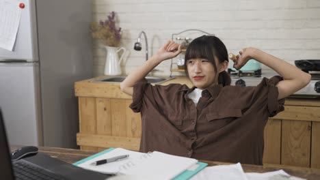 tired korean female employee is leaning back in chair to stretch arms while taking a break from work on the computer in the dining room at home during daytime.