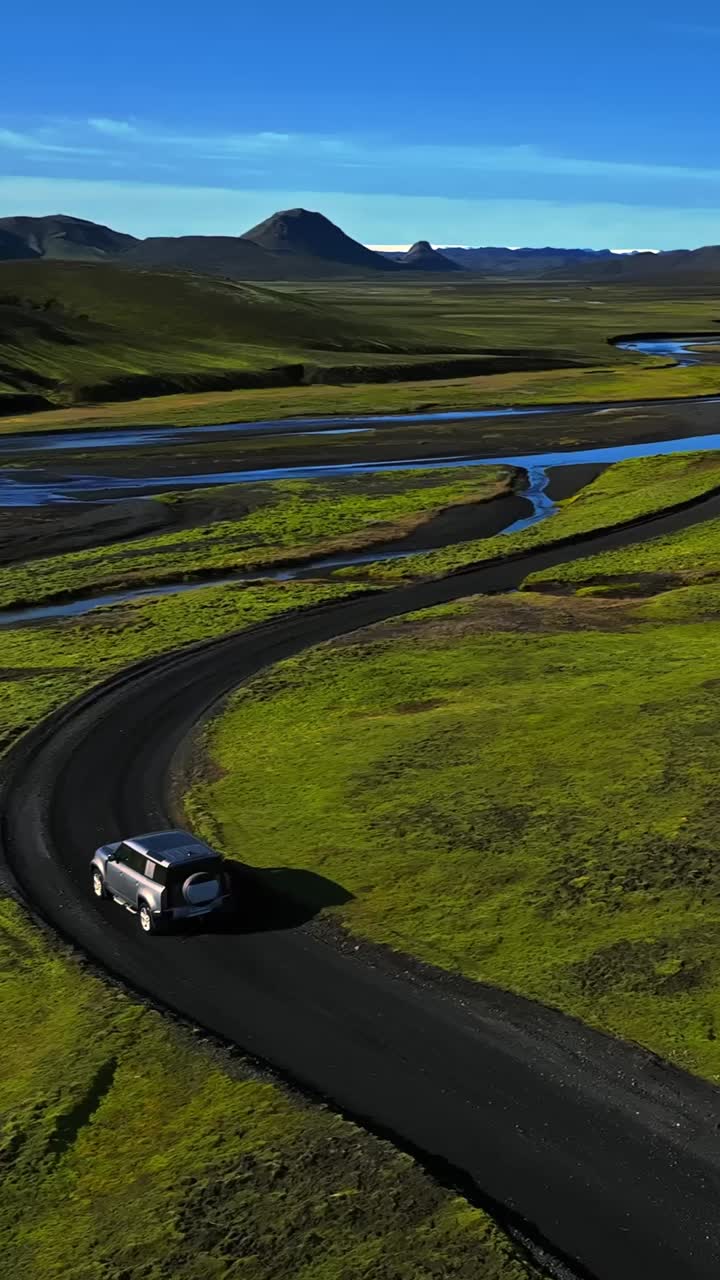 Drone verticale che segue un SUV 4x4 mentre percorre una strada sterrata curva lungo i canali intrecciati di un fiume attraverso muschio verde e sabbia vulcanica nera, con montagne in lontananza e cielo blu