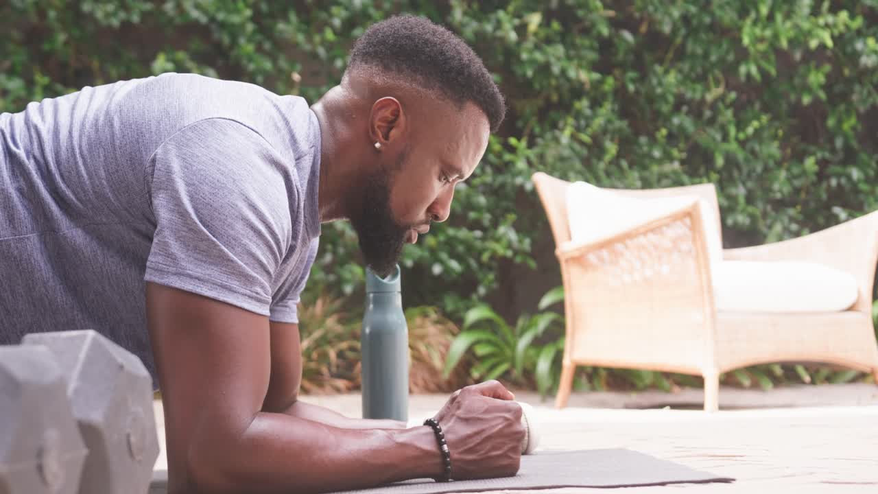 African American man holding forearm plank on mat at patio, wearing grey tee, water bottle nearby