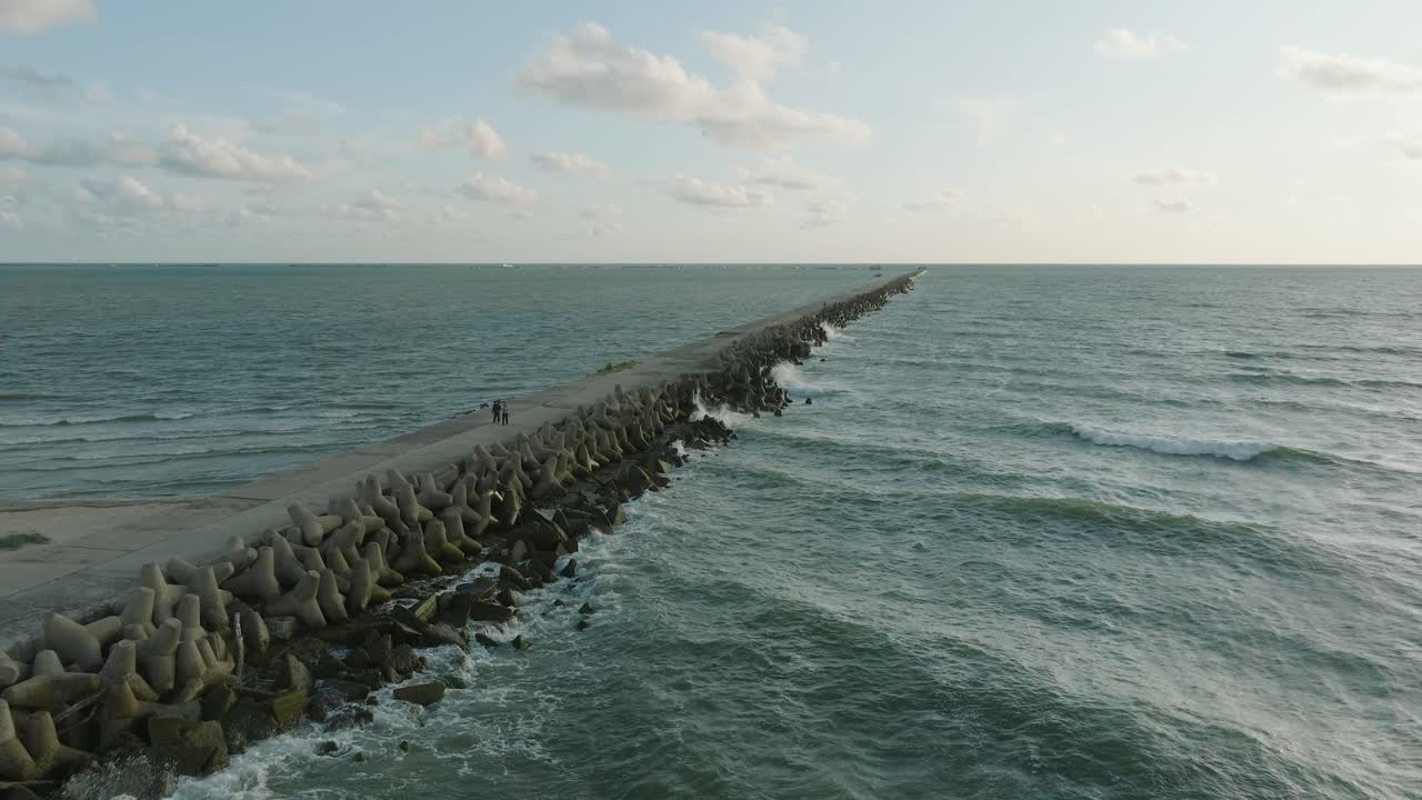 Vista aérea panorámica del muelle de hormigón del Puerto de Liepaja, costa del mar Báltico, tarde de verano soleada, luz de la hora dorada, grandes olas rompiendo, toma amplia de dron avanzando.