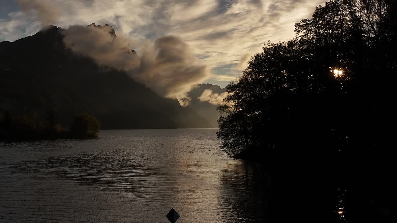 Drone slowly ascends from Walensee lake, majestic mountains with clouds gently touching their peaks during stunning morning sunrise in Switzerland.