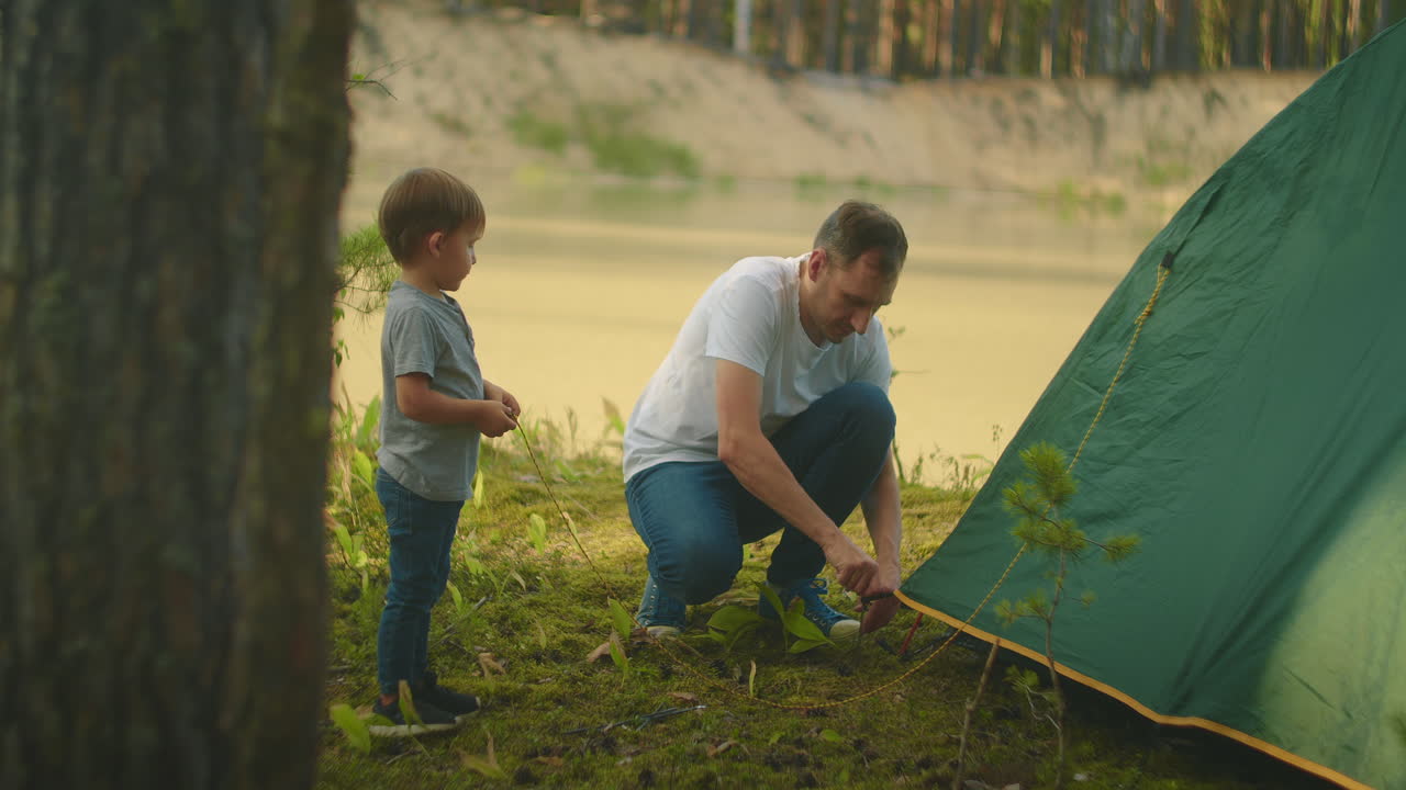 el niño ayuda a su padre a montar una tienda de campaña en la orilla del lago en el bosque