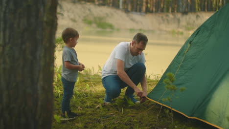 little boy helps his father set up a tent on the shore of the lake in the woods