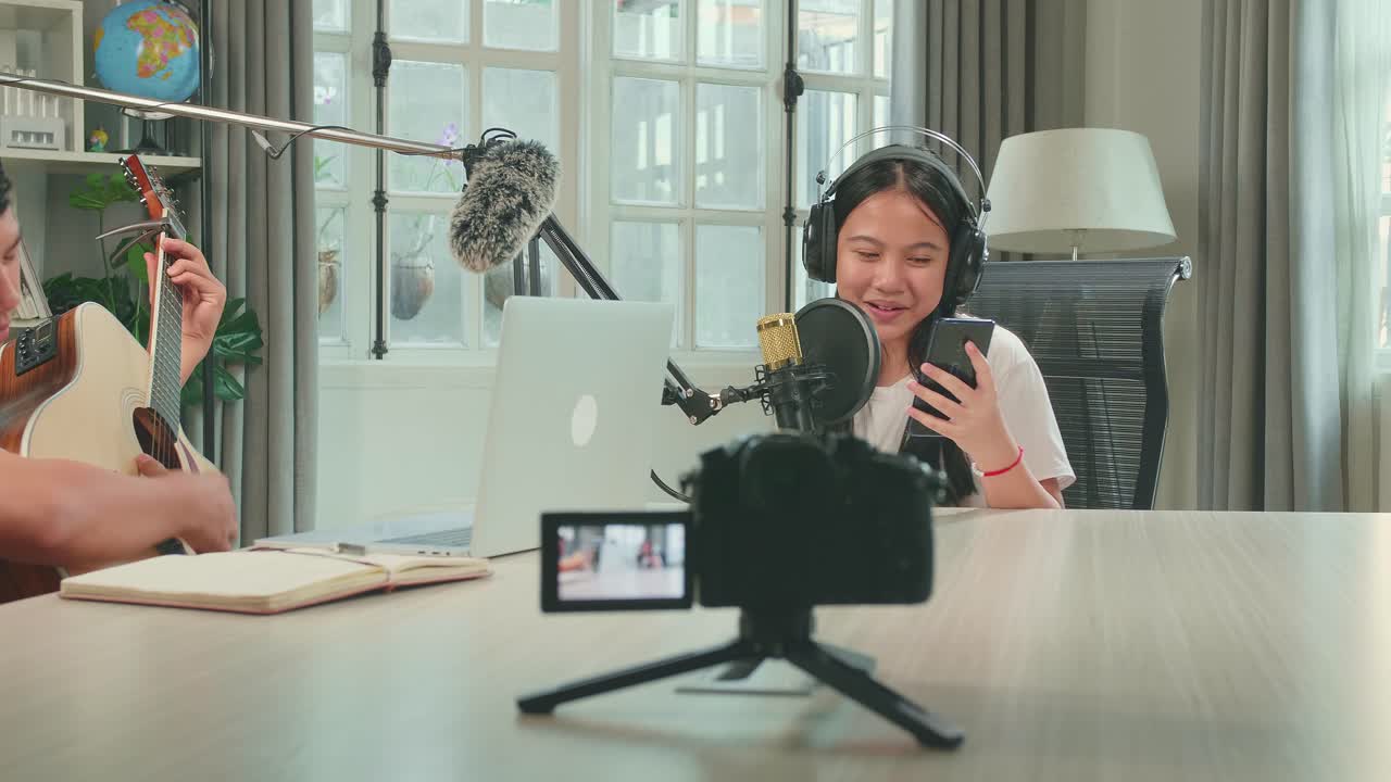 Camera Recording, Asian Kid Girl Holding Phone And Wearing Headphones Singing Into Microphone While Recording Podcast With Boy Playing The Guitar In Studio