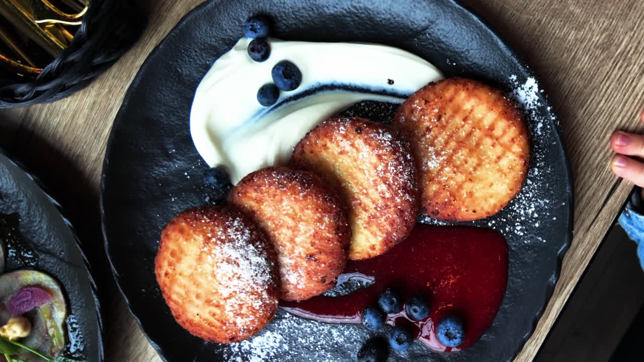 Golden pancakes served with whipped cream, blueberry sauce, and fresh berries on a dark plate, perfect for a rustic breakfast or elegant brunch presentation