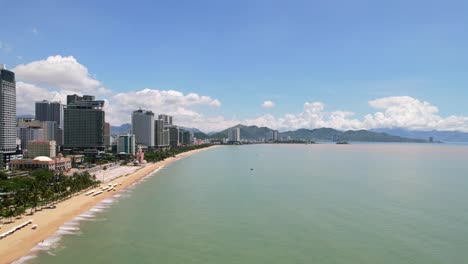 Luftaufnahme-Aus-Geringer-Höhe-Der-Küste-Von-Nha-Trang-Voller-Weißer-Sonnenschirme-An-Einem-Sonnigen-Tag-Mit-Hohen-Hotels-Mit-Blick-Auf-Die-Palmen-Und-Das-Türkisfarbene-Wasser