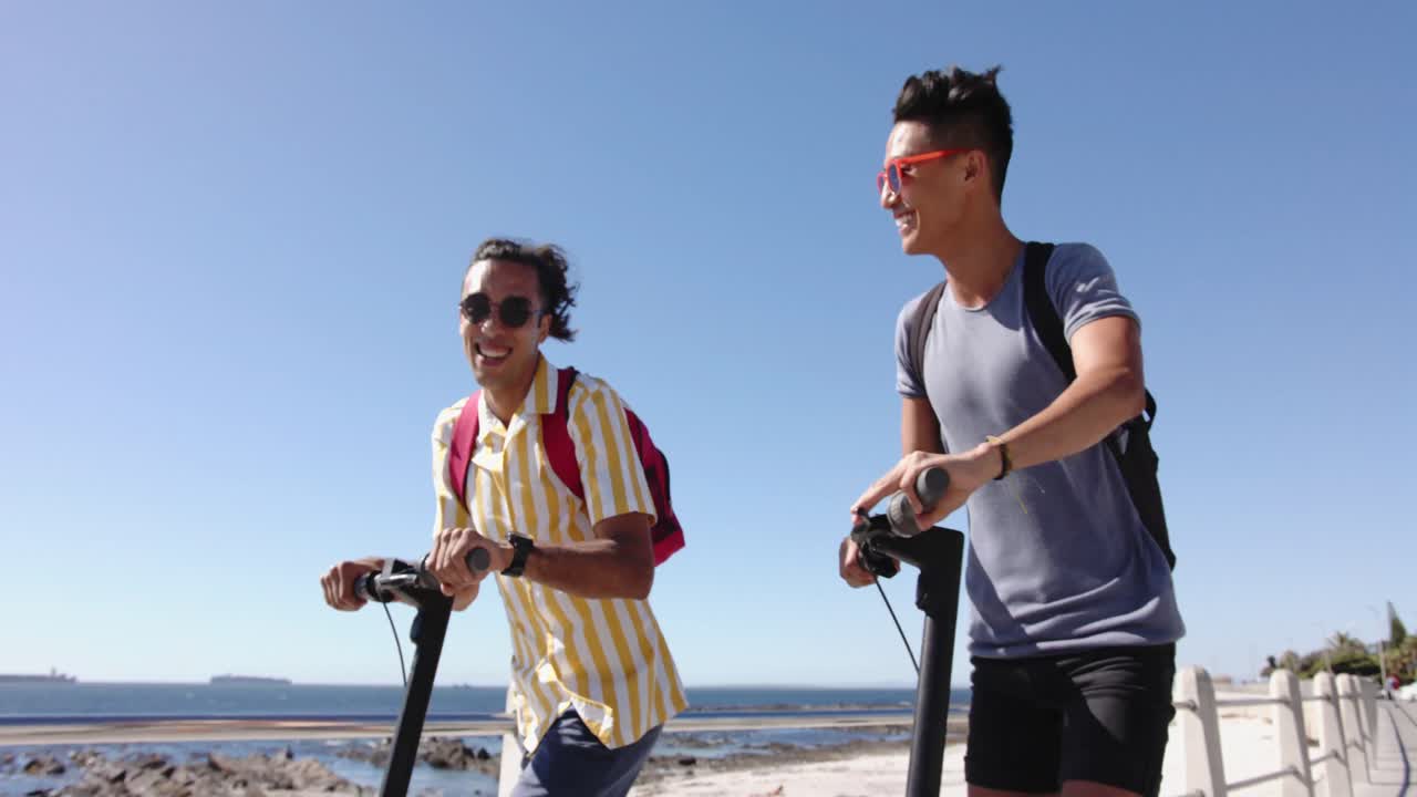 해변의 산책로에서 스쿠터를 사용하는 행복한 다양한 동성애 남성 커플, 슬로우 모션