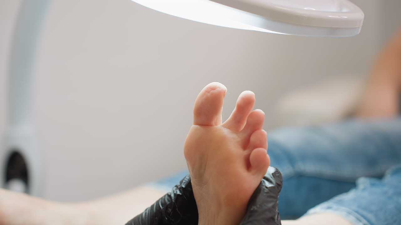 Close-up of technician in black gloves massaging underside of client foot under bright lamp. Foot detail emphasized while background remains softly blurred, highlighting therapeutic care