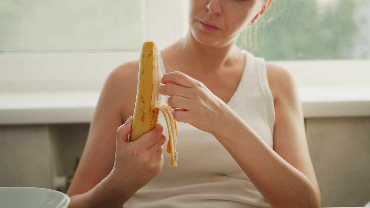 Chef peeling back of banana carefully with both hands near window in bright kitchen, dressed in white sleeveless top, preparing fresh fruit for meal with focused attention in clean indoor environment