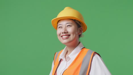 close up side view of asian female engineer wearing safety helmet looking around while standing with arms akimbo in the green screen background studio