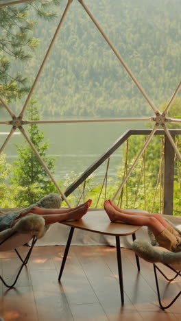 children rest in glamping cabin at lakeside. little boy and girl sit in comfortable chairs under transparent dome. kids enjoy lake view at camp