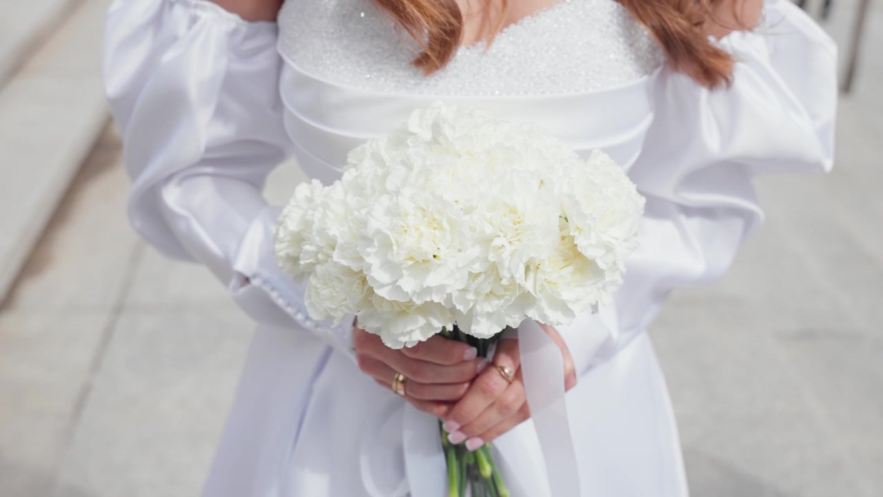 Primo piano romantico di una sposa che tiene un delicato bouquet bianco. L'abito con maniche a sbuffo scintilla alla luce del giorno. L'illuminazione naturale esalta il tessuto elegante, i dettagli floreali e le mani graziose