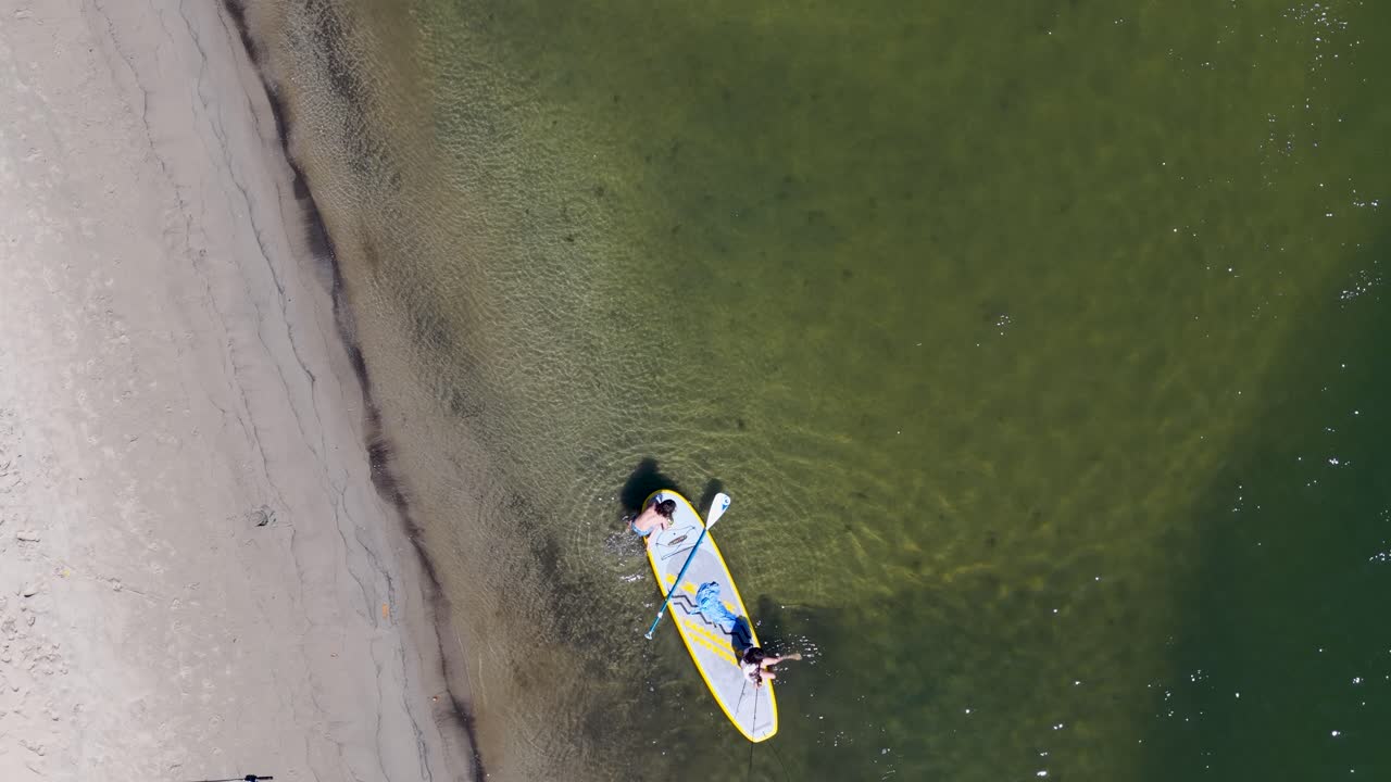 Two children paddleboard and fish in shallow, sunlit water near sandy shoreline, aerial perspective