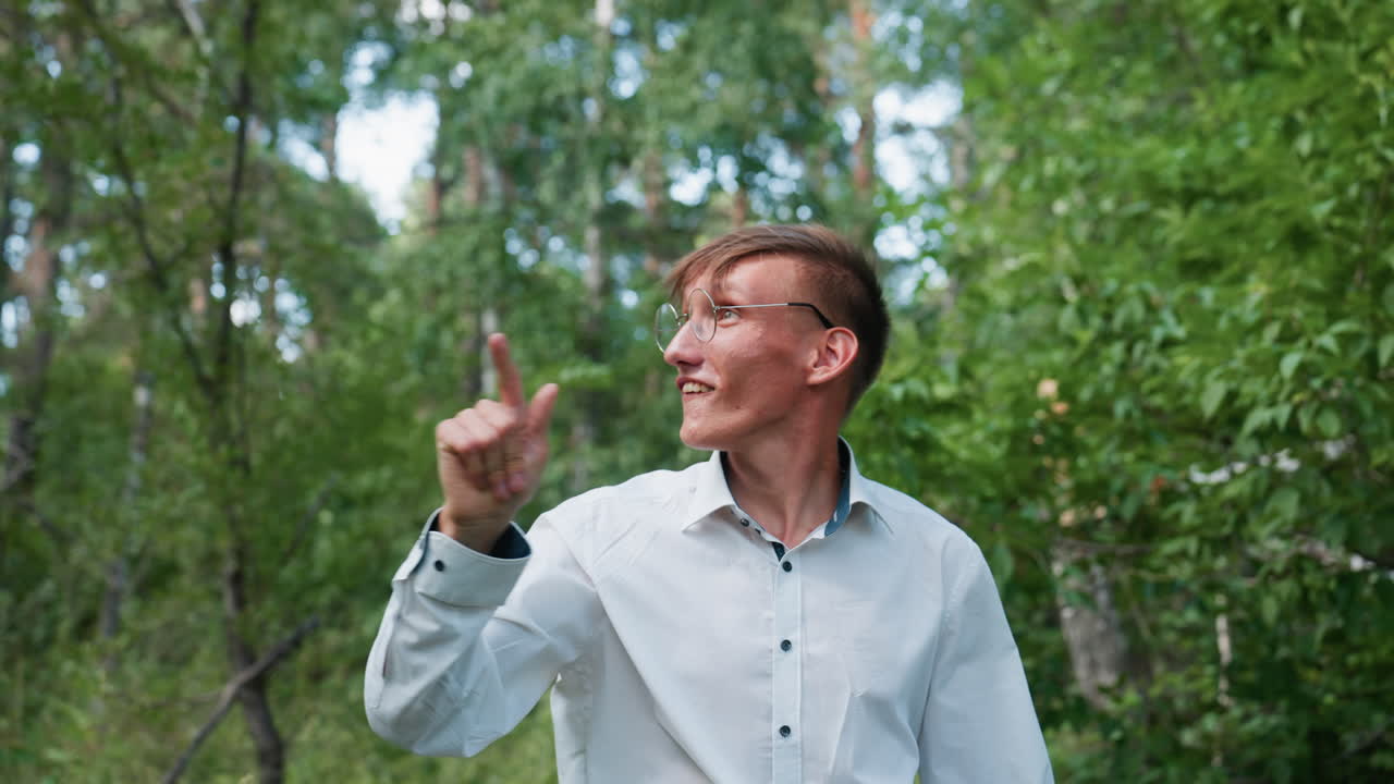 Closeup view of young man in white shirt walking slowly through forest path, pulling up glasses while observing plants along way surrounded by lush greenery and soft natural sunlight