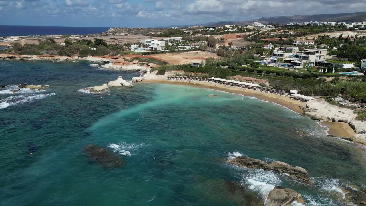 Aerial drone captures Maniki Beach, Cyprus, revealing cinematic views of crystal-clear blue waters, rugged rocks, and stunning coastal homes beautifully set against the serene sea.