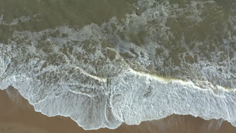 Aerial-view-of-high,-powerful,-white-foam-waves-slowly-crashing-on-an-empty-sandy-beach