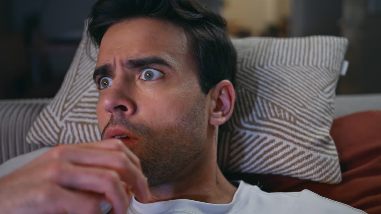 Amazed guy watching thriller on TV home lying sofa at night closeup. Shocked man