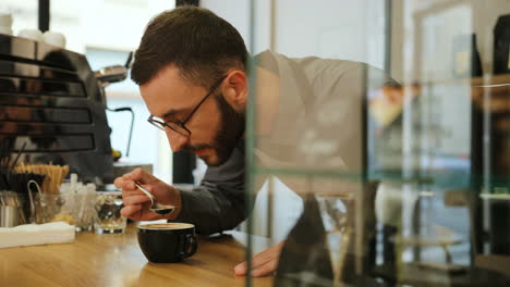 close-up view of caucasian male barista with beard and glasses tasting the fresh coffee in a coffee shop