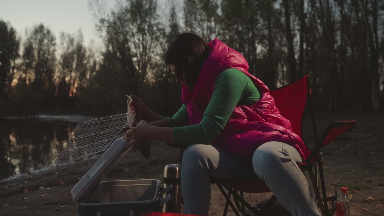 Aftenskumring ved søen, campingkvinde rengør udstyr ved siden af klapstol, skyller fiskenet over transportabelt komfur og køleboks, pink vest og grøn sweater, træsilhuetter ved solnedgang, lanterneglød kaster et varmt skær