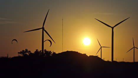 Silhouettes-of-kitesurfers,-palm-trees-and-wind-turbines-during-a-hazy-sunset-in-Brazil