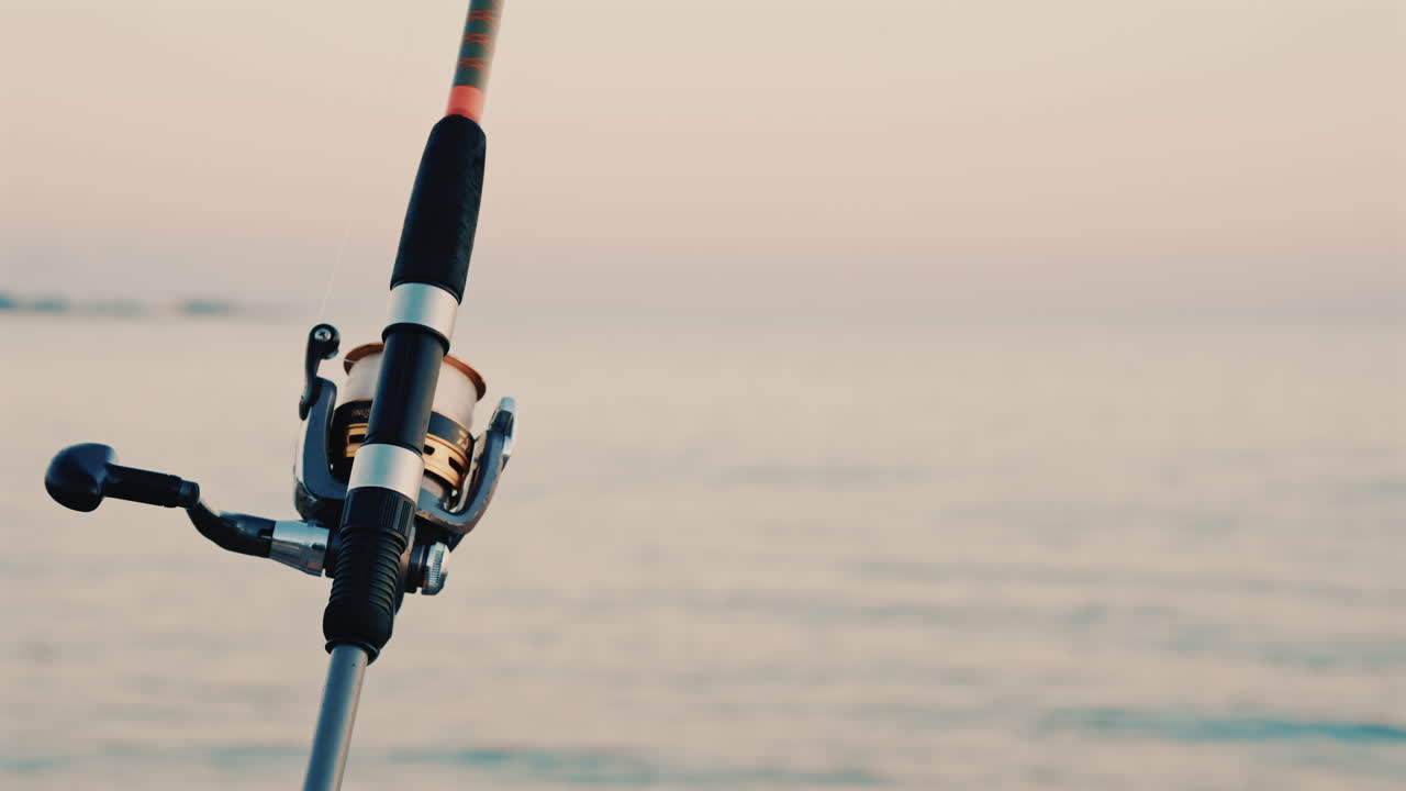 Close up of a fishing rod and reel positioned near the sea, with a blurred boat in the background