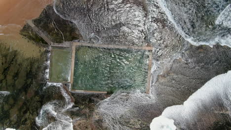 Toma-Aérea-De-Ojo-De-Pájaro-De-Las-Olas-Y-El-Poderoso-Oleaje-Del-Océano-Rompiendo-Sobre-La-Piscina-De-Rocas-De-Monavale-En-Nsw,-Australia-En-Un-Hermoso-Día-De-Primavera