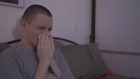 a young man sitting on bed and blowing his nose on a tissue paper- medium shot