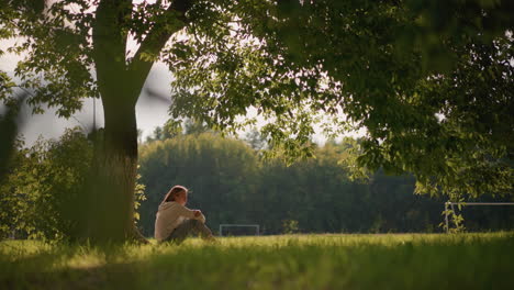 woman sits peacefully by a large tree in lush greenery, sunlight casting a warm glow, background shows distant trees, a football goalpost, and subtle movement of leaves and a figure in the distance