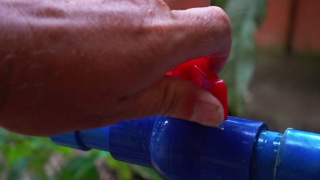 un primer plano que muestra la mano de un adulto girando un grifo rojo en sentido antihorario en una tubería de agua azul de plástico