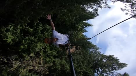 vertical action camera shot of a young man, happily swinging out from the platform up in the trees, strapped to a zipline and lying down and stretching his arms and legs out in freedom