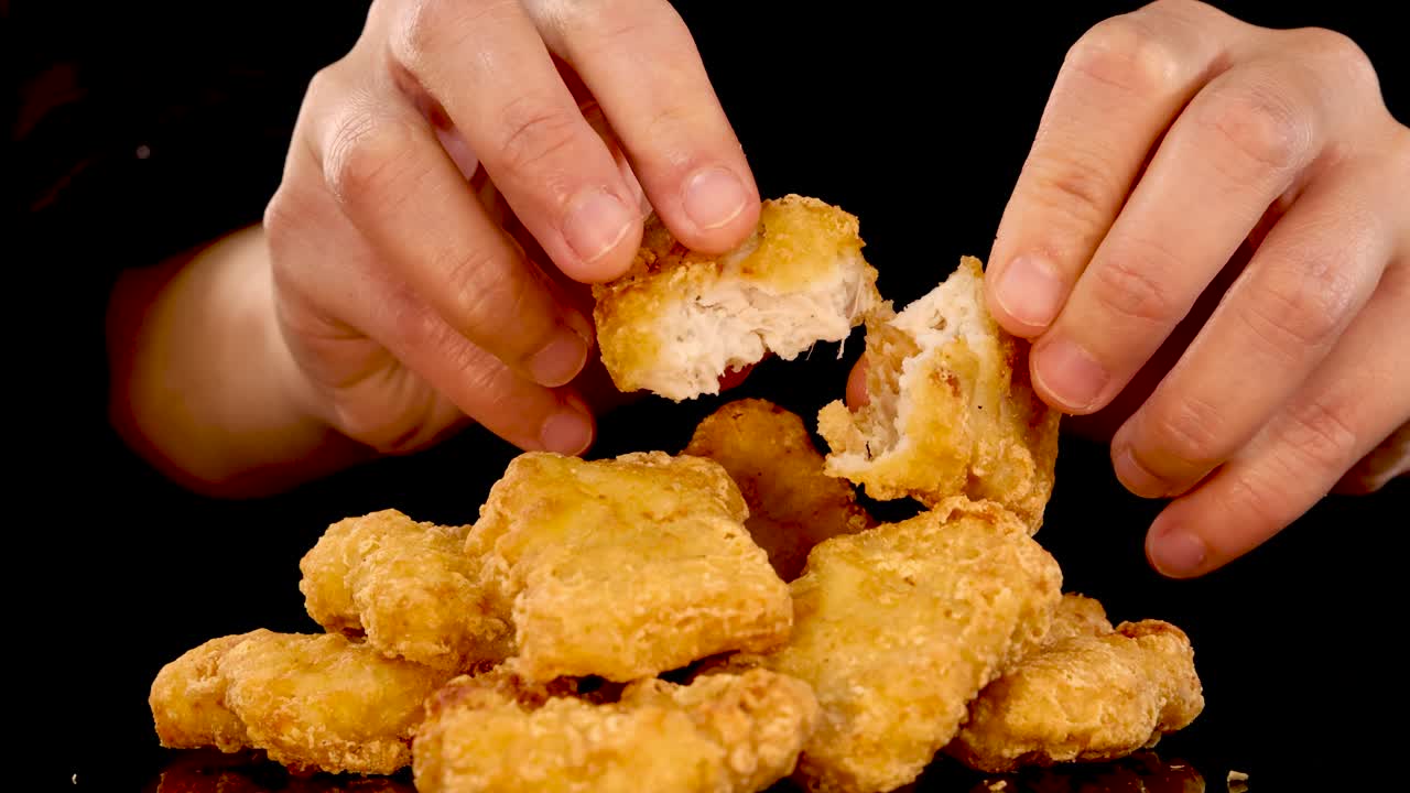 A pair of hands picks up and splits open a spicy, crumbed fried chicken nugget over a pile, under dramatic studio lighting with a black background