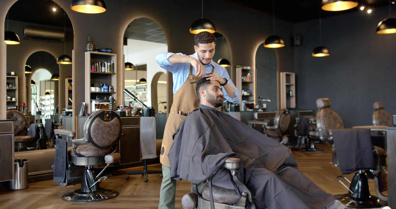 Afroamerikansk barber i forklæde klipper kundens hår i salonen og tjekker for at afslutte med kam og saks.