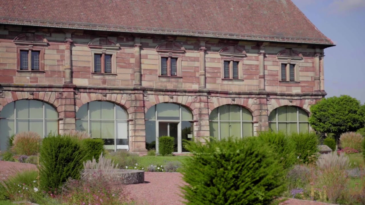 Zeitlupen-Nahaufnahme der Orangerie Blieskastel im Sommer. Betont die filigrane Steinarchitektur, umrahmt von leuchtend grünen Sträuchern und üppiger Gartenumgebung