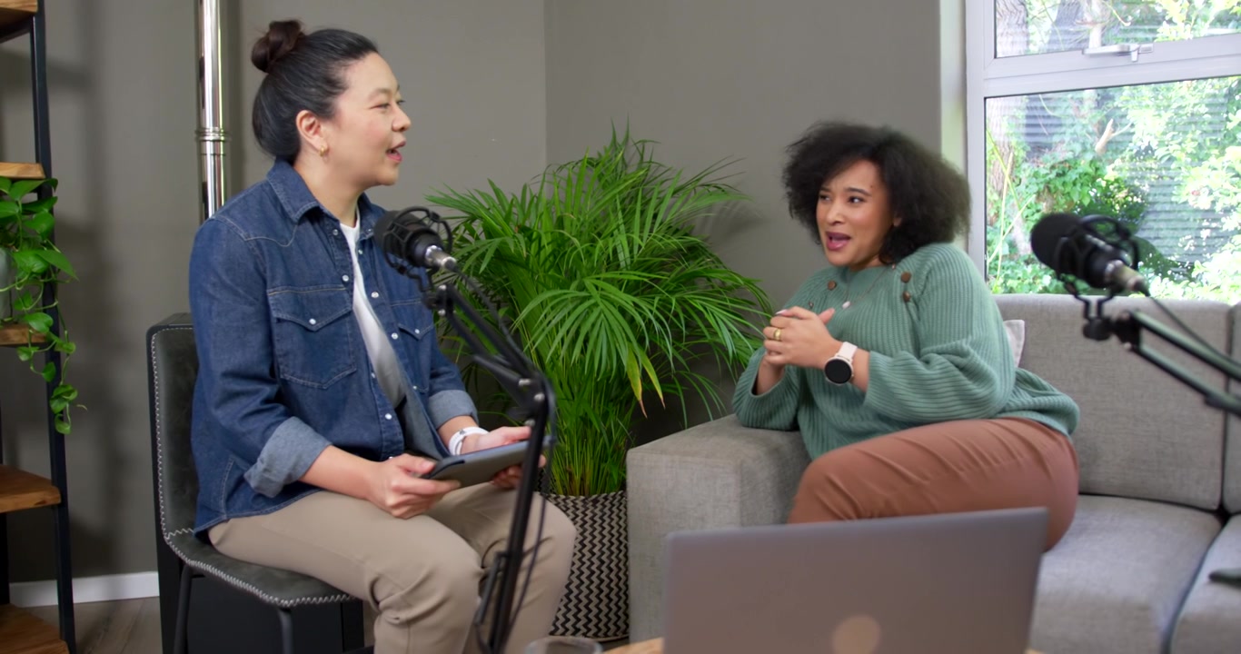 Asian host denim checking tablet, African American guest sage sweater gesturing into podcast mics