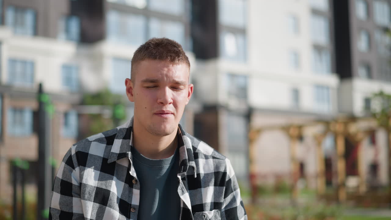 Portrait view of sick boy in checkered shirt holding pill close to mouth, eyes closed in discomfort while standing outdoors under bright sunlight with modern residential buildings in background
