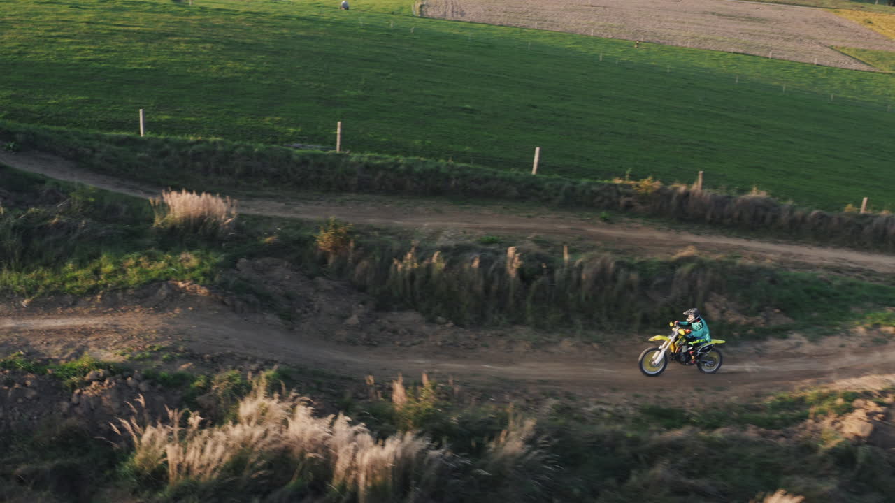 fotografía aérea de un piloto de motocross en una bicicleta de tierra amarilla montando rápido en una larga recta en un sinuoso sendero de tierra rodeado de hierba verde y campos.