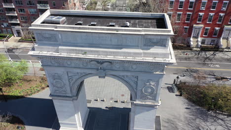An aerial view of the Washington Square Arch in NYC. The drone camera dollys in - tilts down over to the top of the arch. The park - the surrounding streets are empty - it's a bright - sunny day.