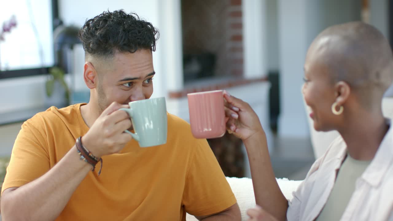 Coppia afroamericana seduta sul divano, la donna alza una tazza rosa invitando a brindare, l'uomo fa tintinnare una tazza color menta.