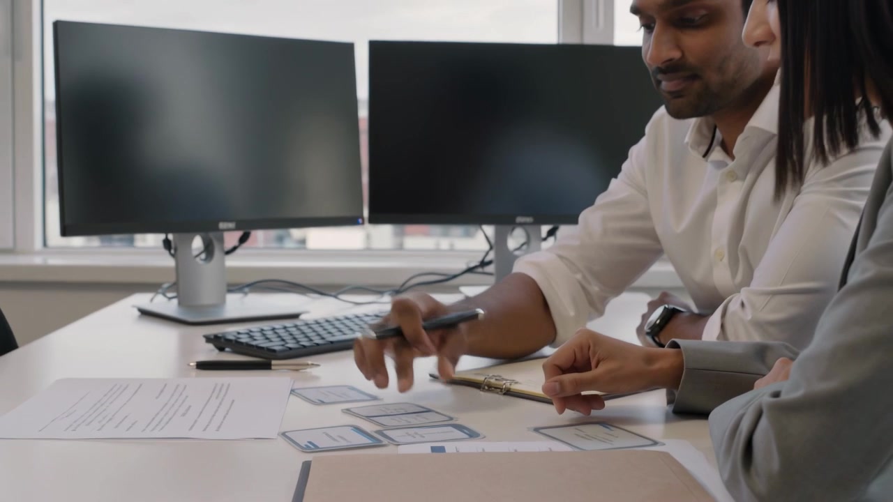 Tapping pen on mobile-wireframe card, man shirt woman blazer pointing and planning layout at desk