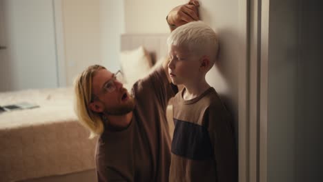 Ein-Blonder-Junge-Mit-Brille-Und-Bart-Misst-Die-Größe-Seines-Kleinen-Albino-Jungen-Mit-Weißem-Haar,-Der-An-Der-Wand-Steht-Und-Darauf-Wartet,-Dass-Sein-Vater-Ihm-Seine-Größe-Sagt