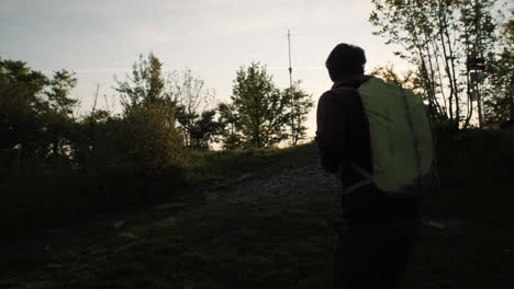 hiker with a green backpack walks up a hill towards the sun, camera tracking from behind