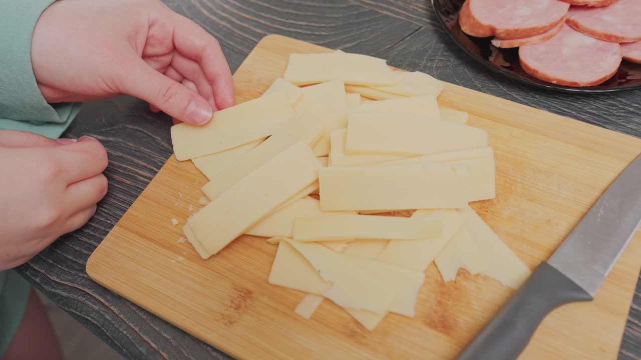 close up of woman in green shirt holding stack of yellow cheese slices above wooden cutting board beside black plate filled with sausage rounds and kitchen knife on counter near electric kettle