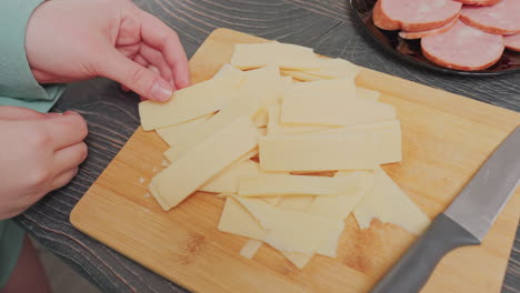close up of woman in green shirt holding stack of yellow cheese slices above wooden cutting board beside black plate filled with sausage rounds and kitchen knife on counter near electric kettle