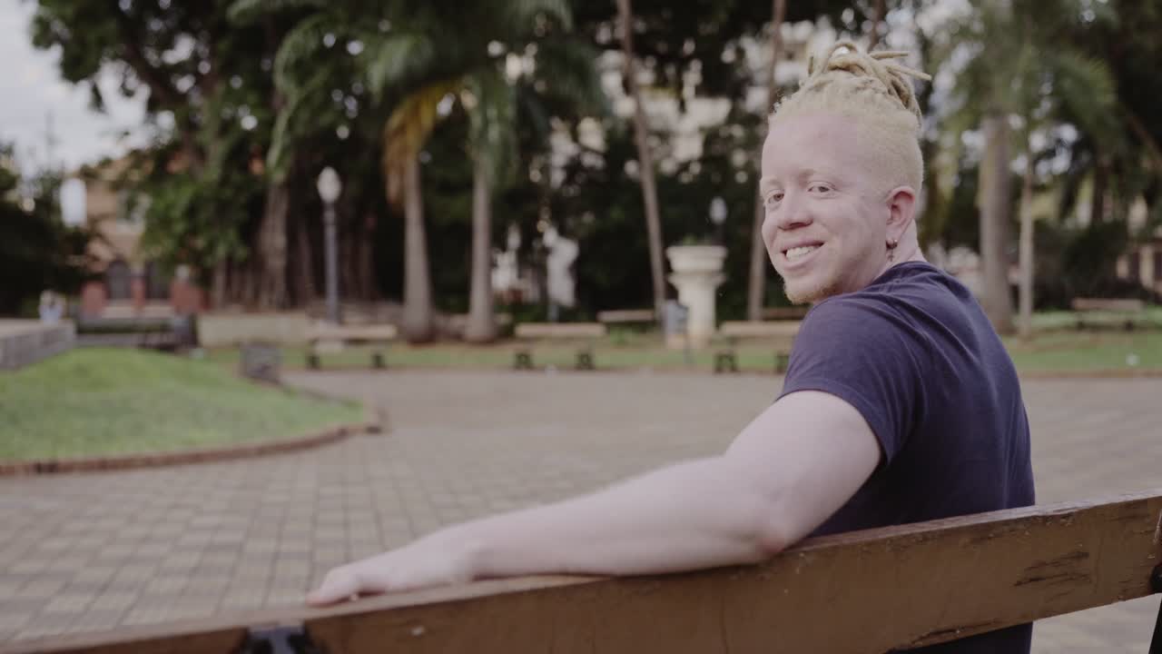 A smiling man with albinism and dreadlocks sitting on a park bench