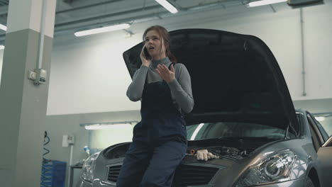 Mechanic female using a smartphone having a call in a car workshop. International Women&#039;s Day.