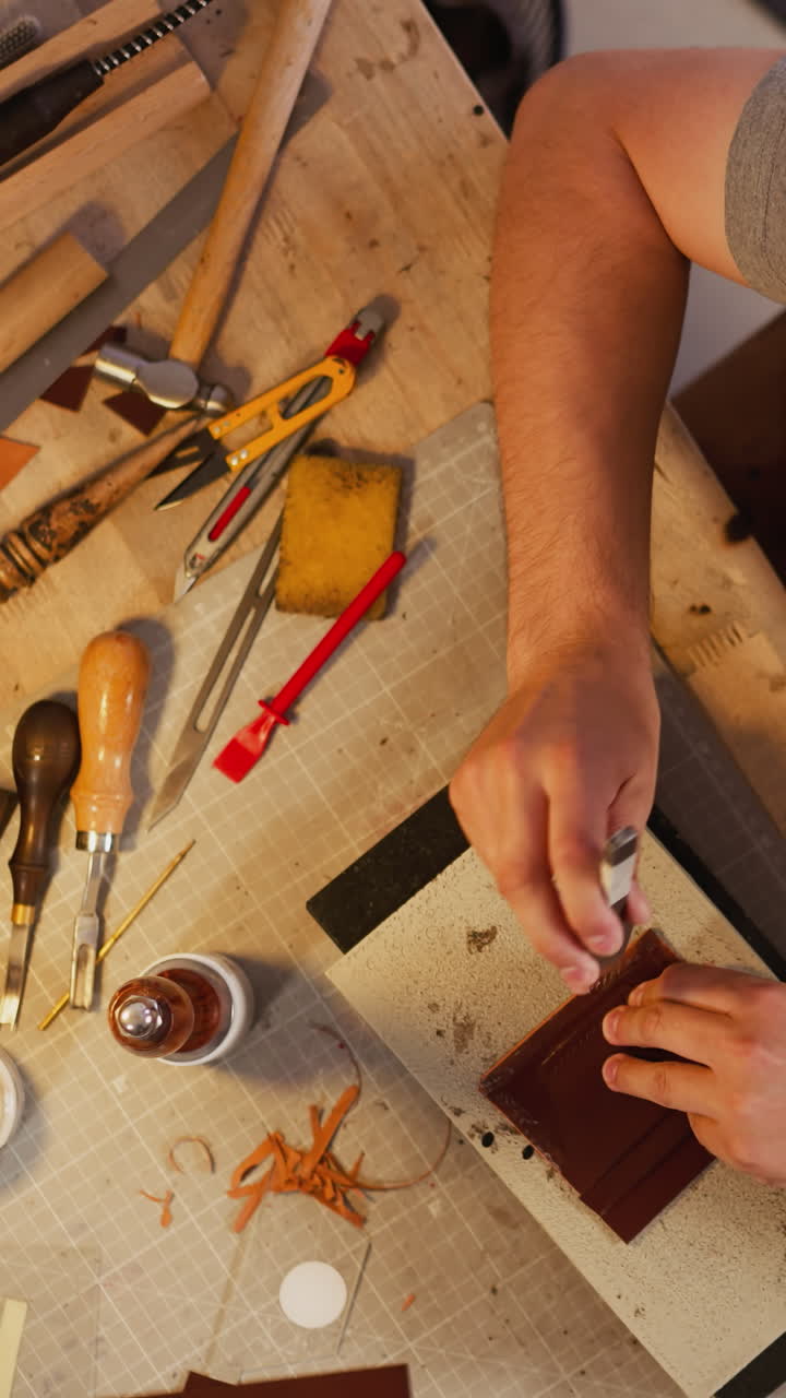 Craftsman makes holes on leather sheet with punch and special hammer at table in workshop upper view. Tradition of handicraft. Manual production